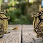 Dos medallas de danza doradas «Golden Bee» con figuras de bailarinas y motivos de abeja en relieve, sobre una mesa de jardín rústica con cintas azules.